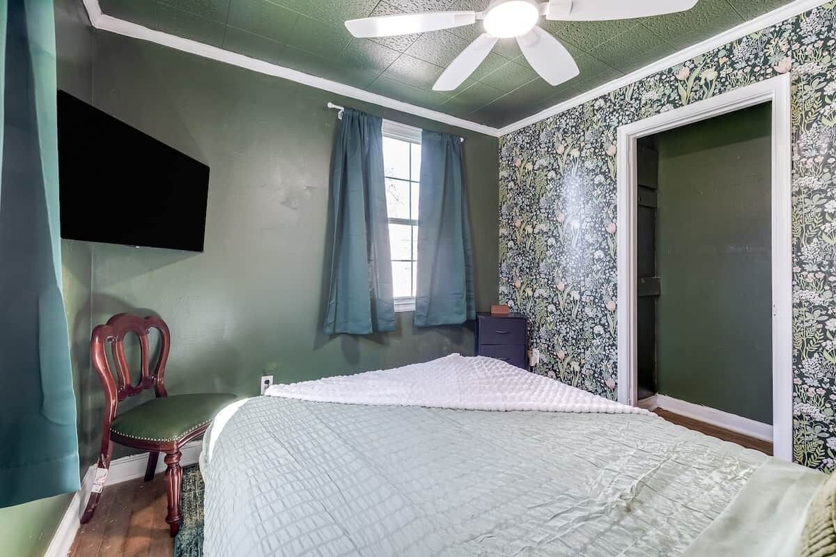 A bedroom with a bed, a chair, a ceiling fan, a window with curtains, and a door.