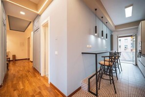 A modern interior with a wooden floor hallway, a kitchen area with bar stools, and a view through a glass door.