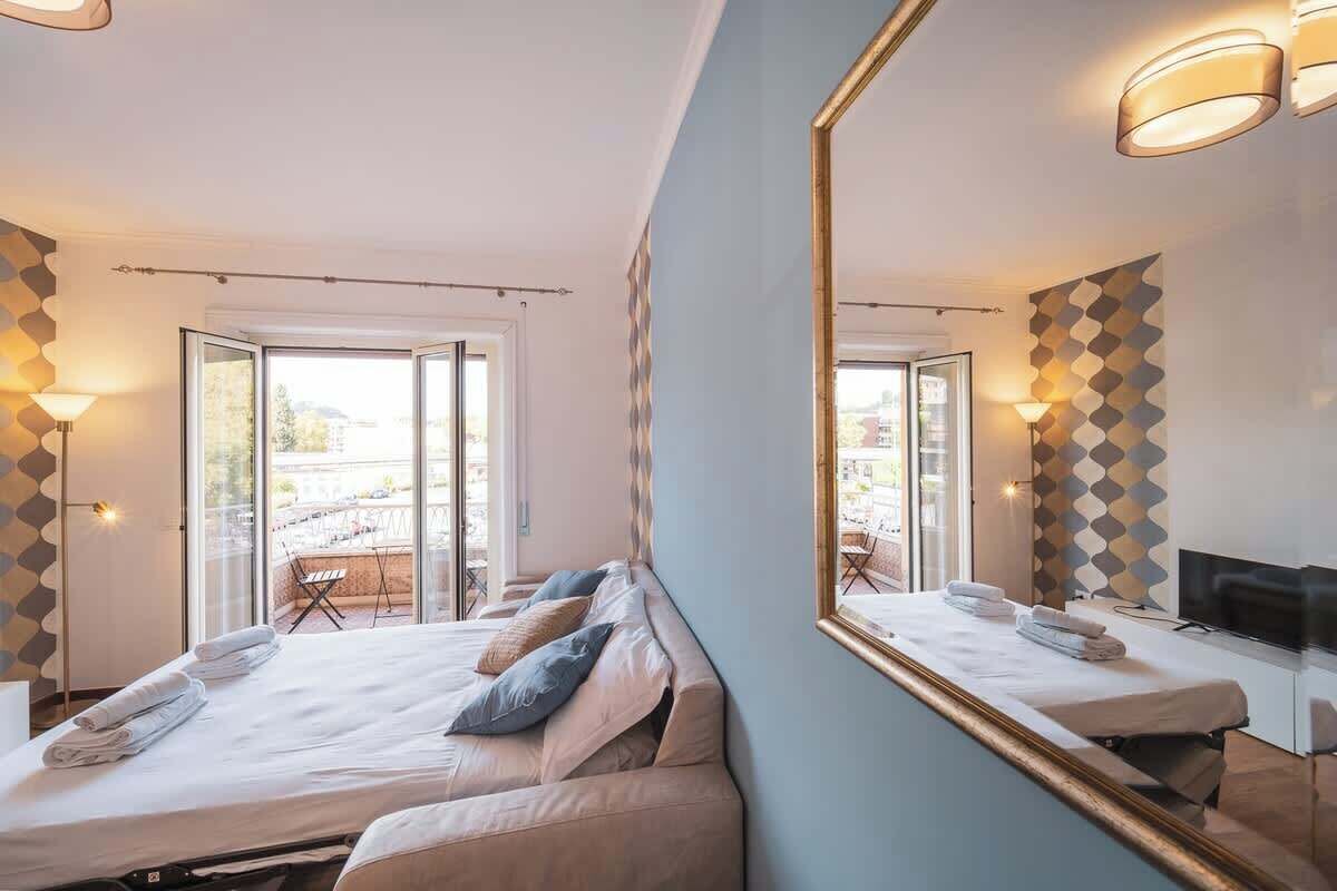 A hotel room with a bed, a TV, a large mirror, and a balcony with outdoor seating.