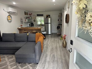 A modern living room with a grey sectional sofa, a wooden dining table with chairs, a kitchen area with a fridge and stove, and a large window with a view. - Cool Cottage near Ennis (Rice)