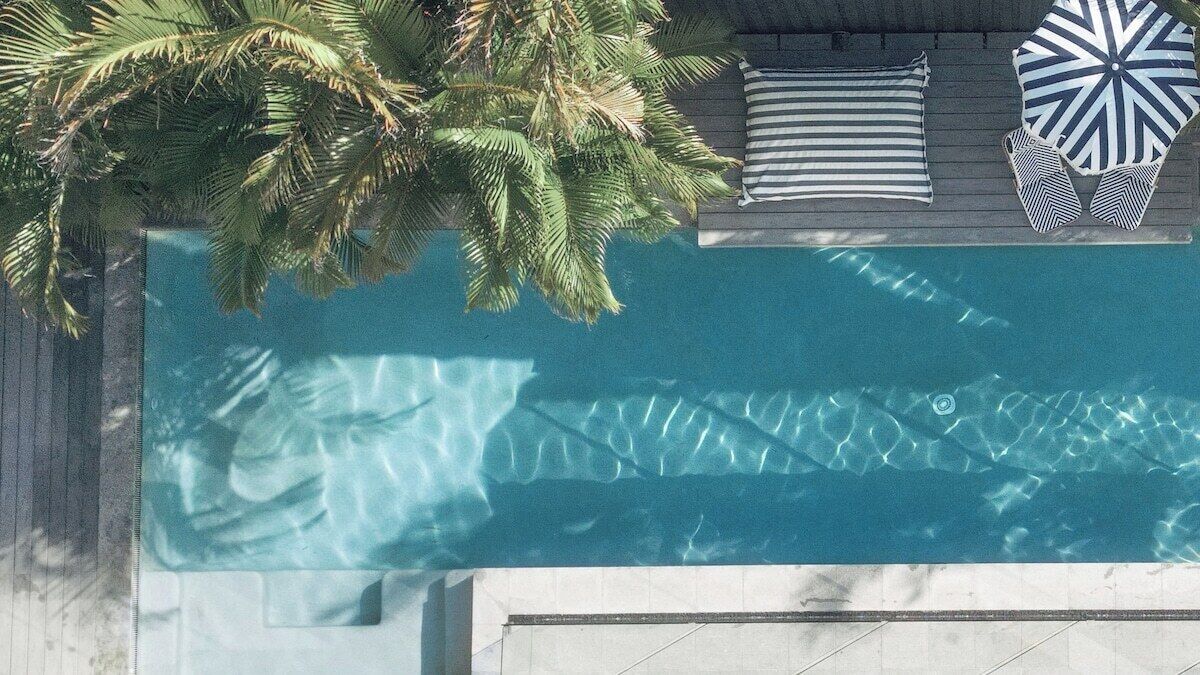 A swimming pool with a wooden deck, a lounge area with striped pillows and a patterned umbrella, and a palm tree.
