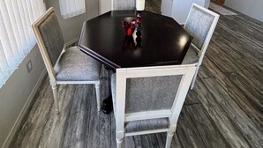 Un coin repas avec une table noire et des chaises blanches, une cuisine en arrière-plan et un panneau sur le mur.
