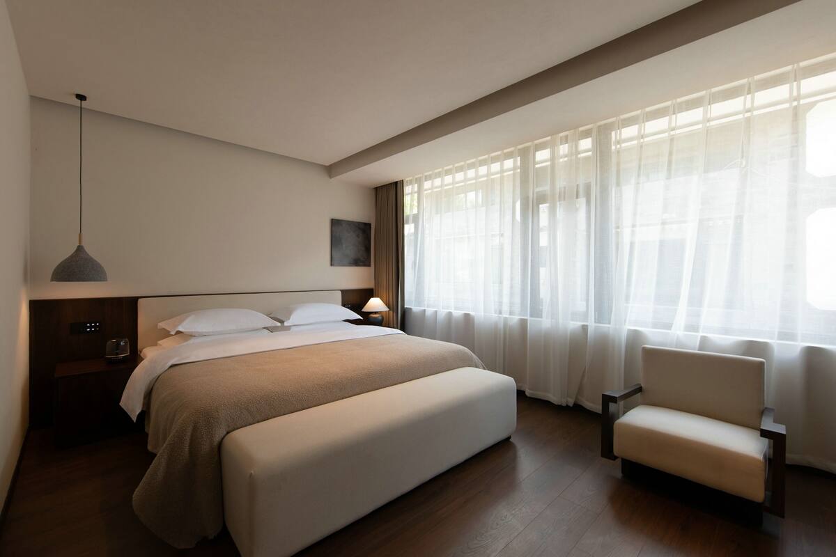 Una habitación de hotel moderna con una cama grande, una mesita de noche con lámpara, una silla y un ventanal con cortinas ligeras.