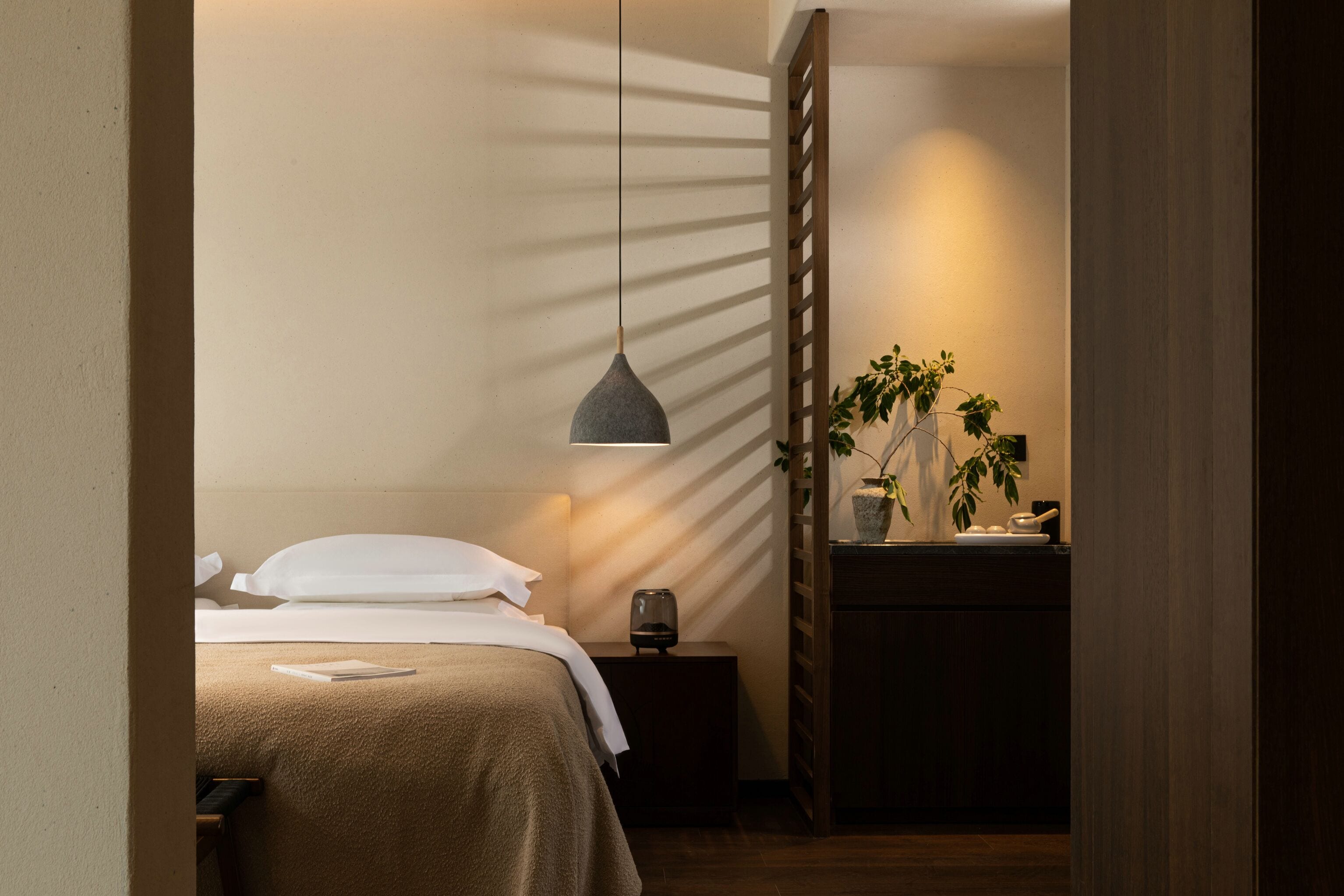 A neatly made bed with a bedside table, a hanging pendant light, and a potted plant on a wooden nightstand.