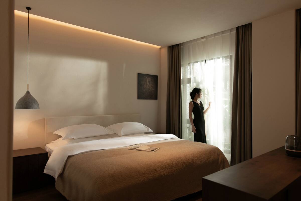 Habitación de hotel con cama, mesita de noche, lámpara y vistas al exterior a través de la ventana.