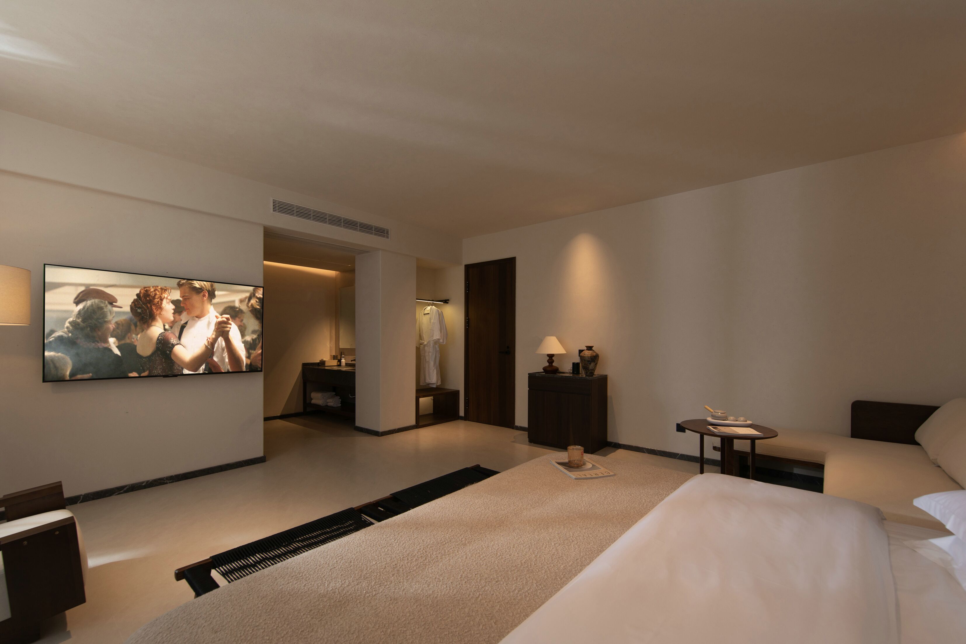 A modern hotel room with a large bed, a seating area, a TV, and a small table with a lamp.