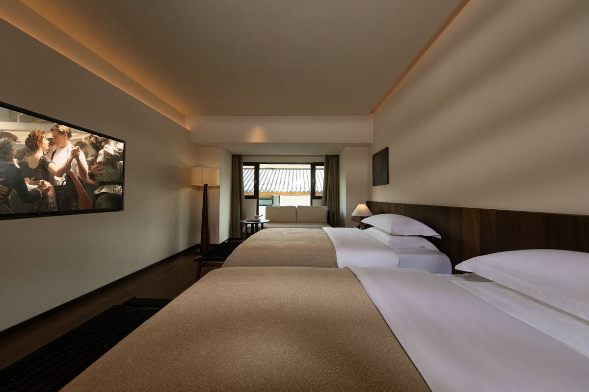 Habitación de hotel con dos camas, un ventanal grande, un cuadro en la pared y un escritorio con lámpara.
