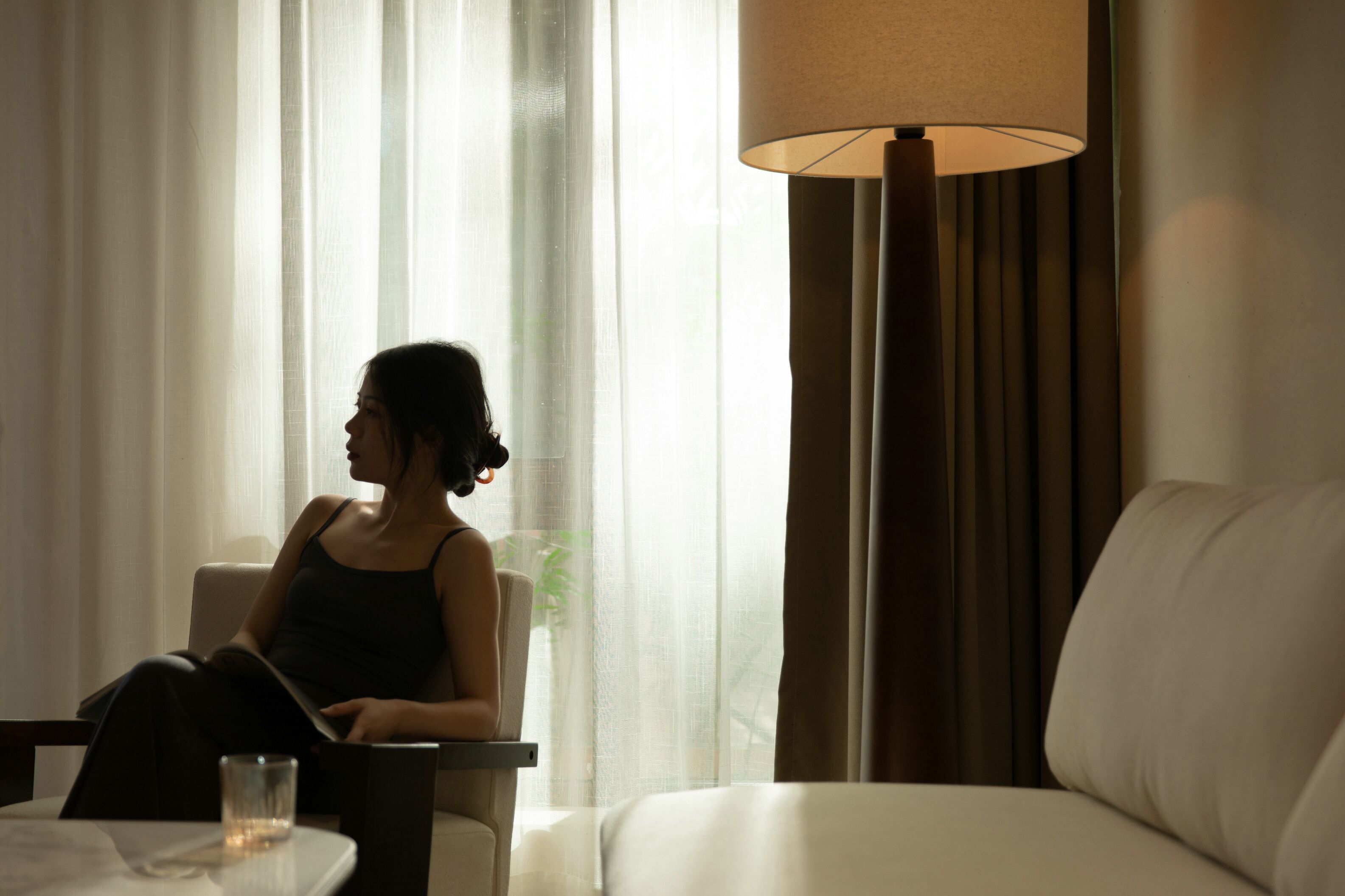 A woman sitting by a window in a hotel room, with a lamp and a glass of water on the table.