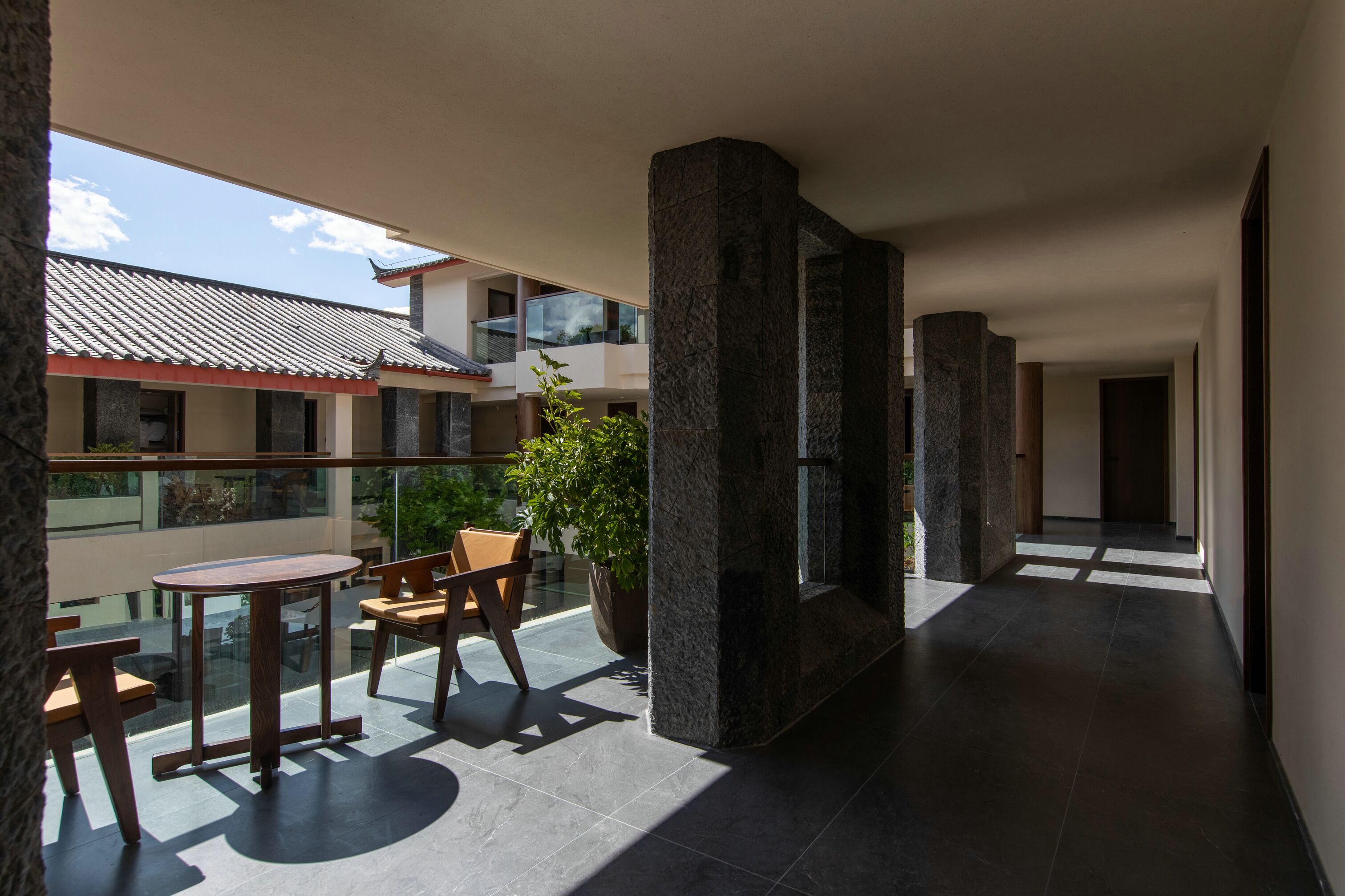 A modern hotel corridor with a glass balustrade, stone pillars, and a seating area with a table and chairs.