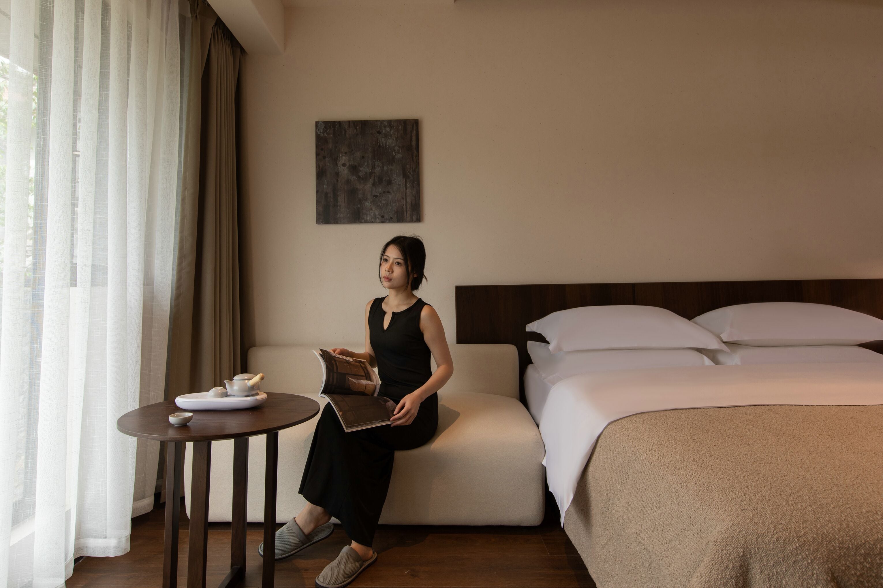 A woman sitting on a bed in a hotel room, reading a book. There is a small table with a teapot and cups, and a window with curtains.