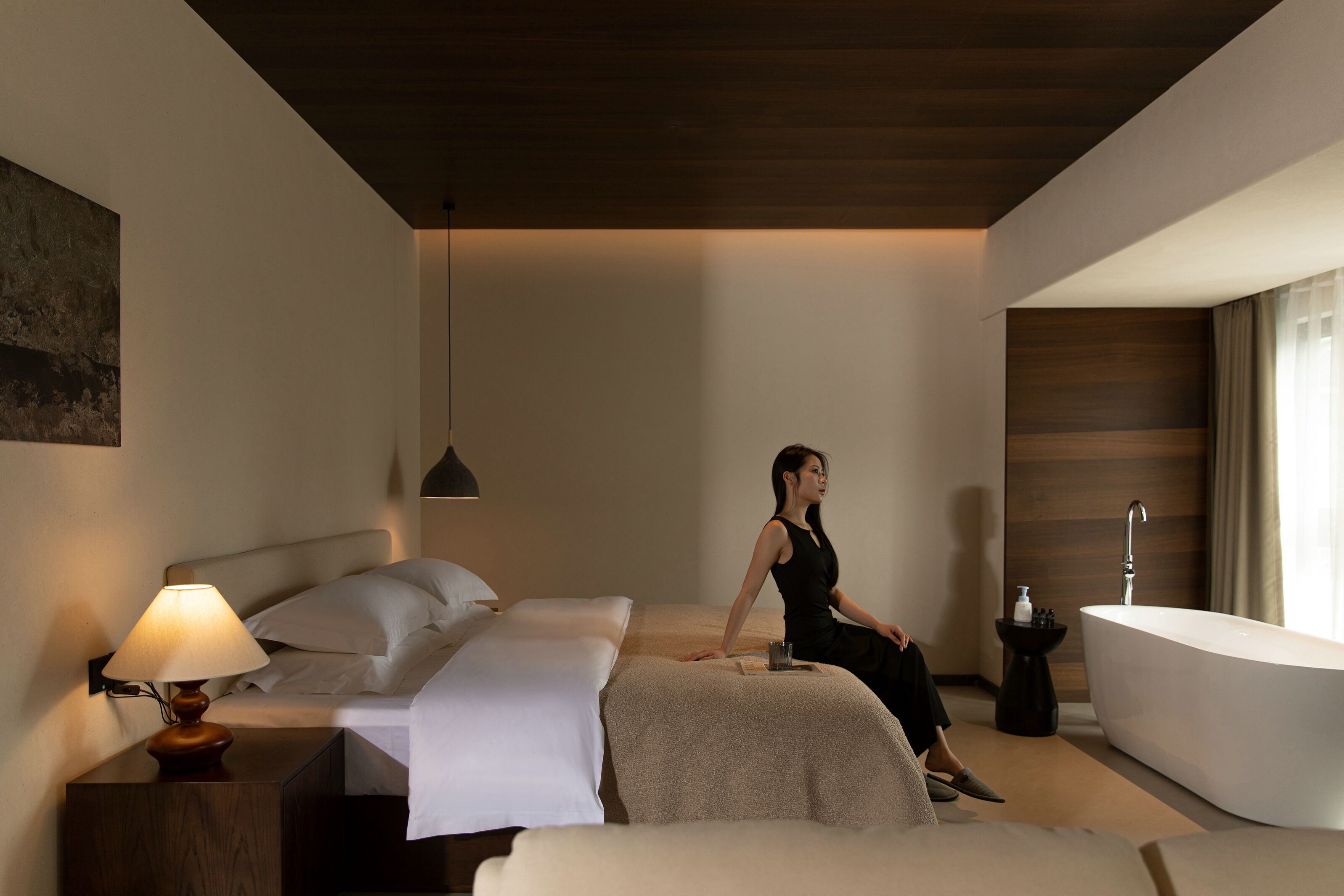 A modern hotel room with a bed, a sitting area, a bathtub, and a nightstand with a lamp.