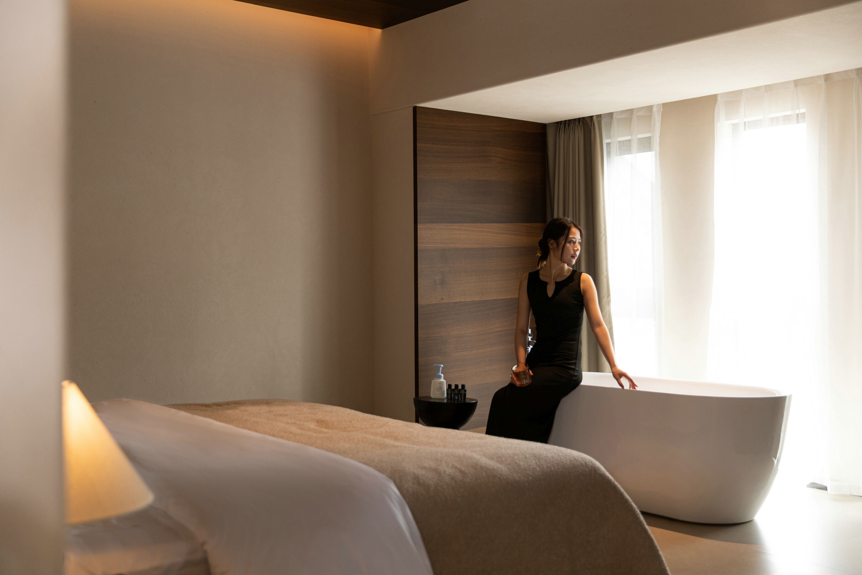 A modern hotel room with a large bathtub, a bed with a white headboard, and a small side table with toiletries.