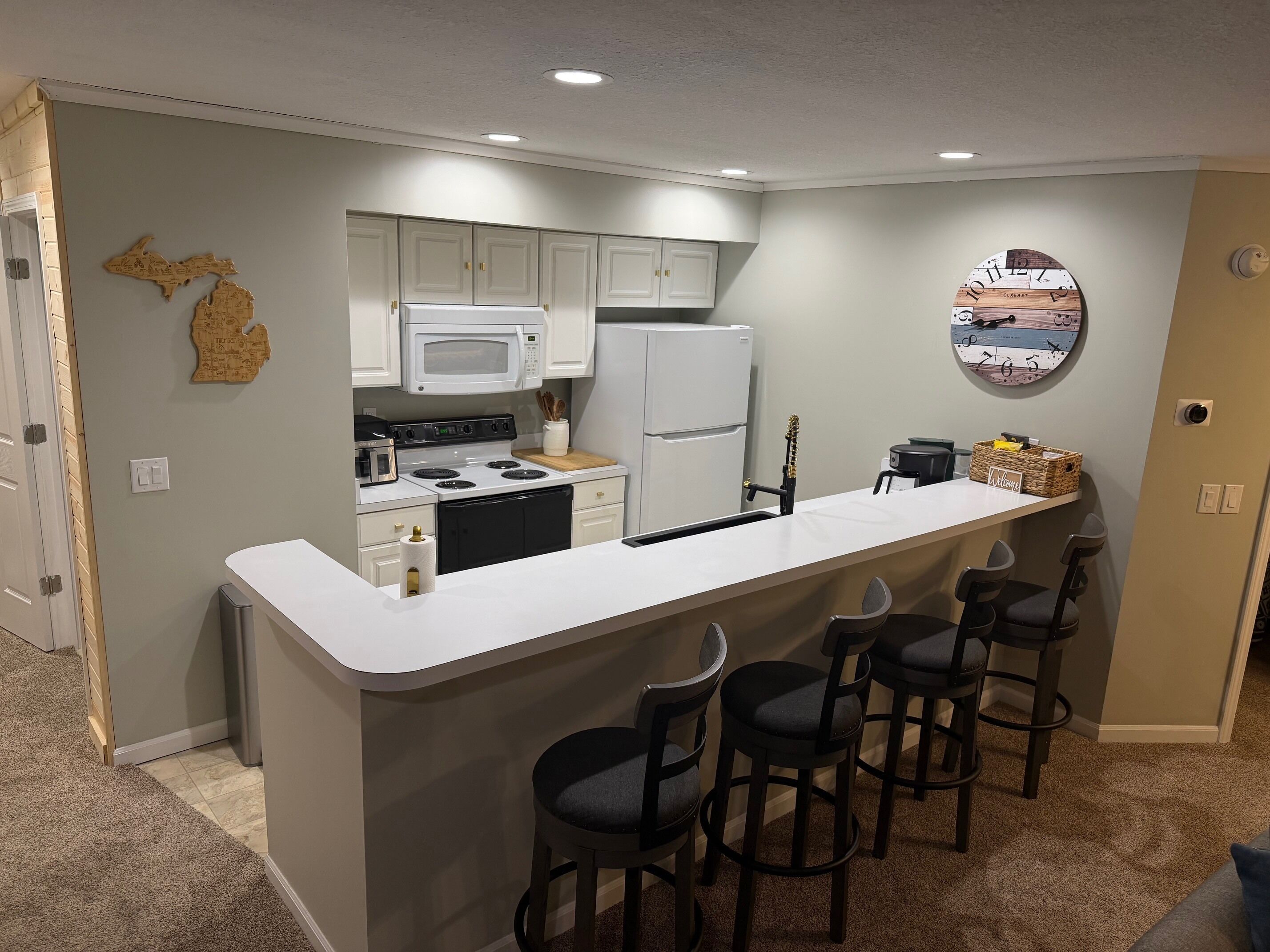 A modern kitchen with a central island, bar stools, a microwave, refrigerator, and a decorative wall clock.
