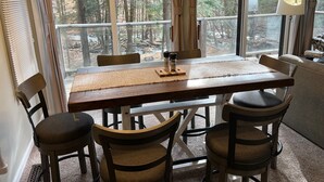Comedor con mesa de madera, cuatro sillas y vista a un bosque a través de amplias ventanas.