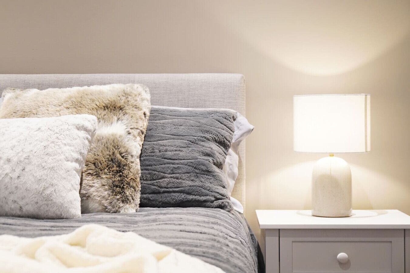 A neatly made bed with a grey and white duvet, a fluffy white pillow, and a grey quilted pillow. There is a white bedside table with a lamp and a drawer.