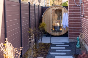 A wooden barrel structure with a bench and a towel hanging on the door, surrounded by a brick wall and a metal fence.