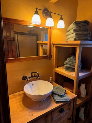 Ein Badezimmer mit einem Holzkosmetikschrank, einem weißen Waschbecken und einem Spiegel darüber. Drei Leuchten sind über dem Spiegel angebracht und auf dem Schrank liegt ein Stapel Handtücher.