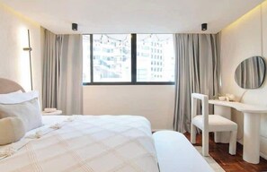 A modern hotel room with a large bed, a desk, and a chair, a window with curtains, and a view of buildings outside.