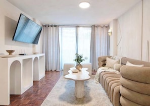 Ein modernes Wohnzimmer mit Flachbildfernseher, weißem Konsolentisch, beiger Couch und großem Fenster mit Vorhängen.