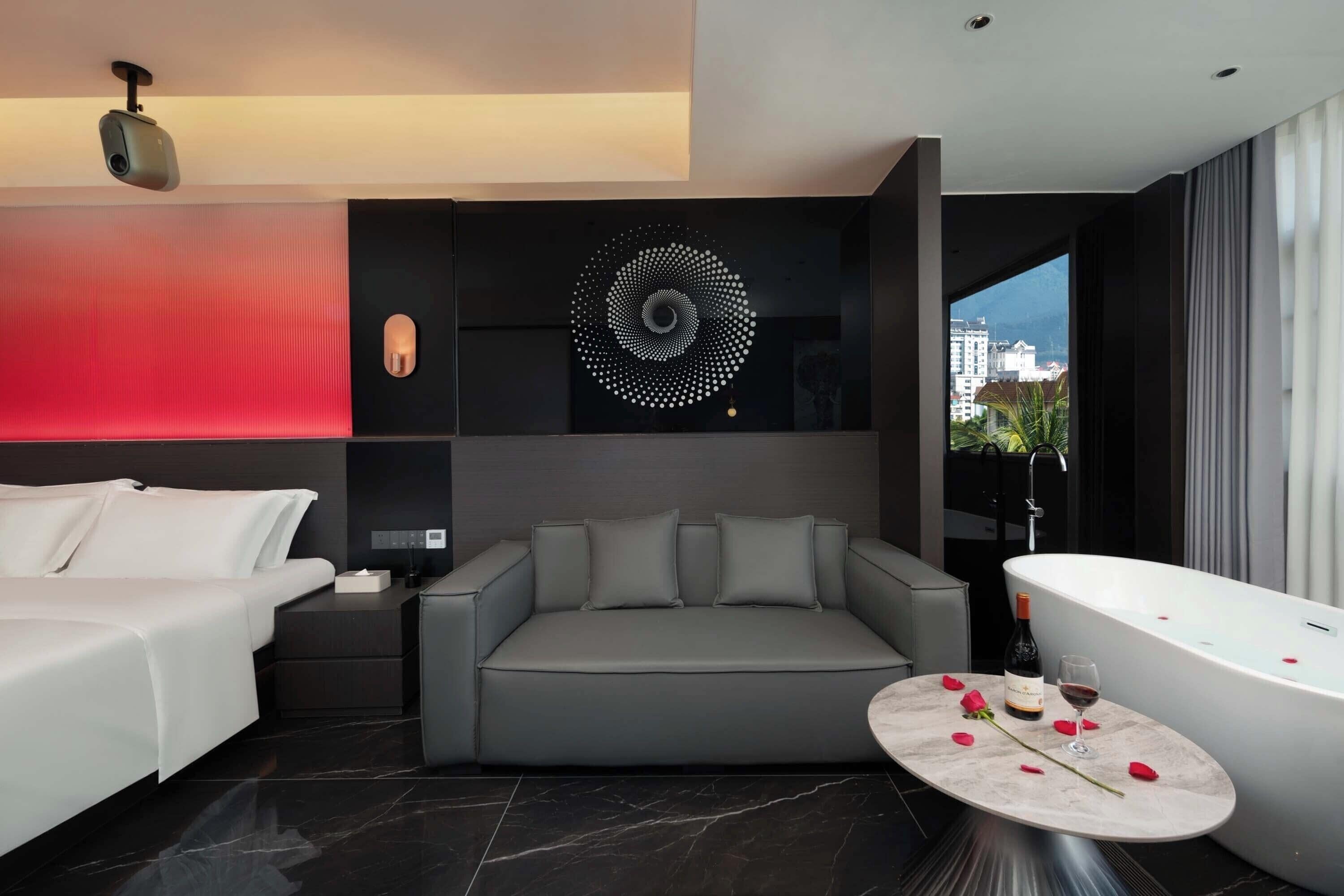 Une chambre d’hôtel moderne avec un canapé gris, un lit blanc, une table ronde en marbre décorée de pétales de rose, une bouteille de vin et une baignoire.