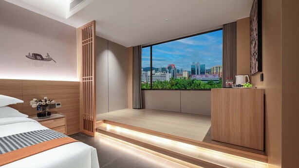 Una habitación de hotel moderna con una cama, mesita de noche y vistas a la ciudad a través de grandes ventanales.