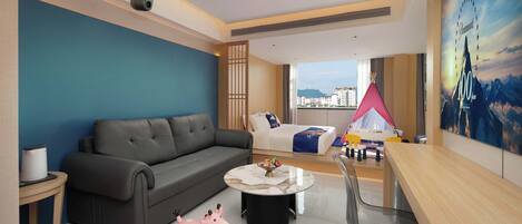 A modern hotel room with a grey sofa, a round coffee table, a desk, and a view of the city.