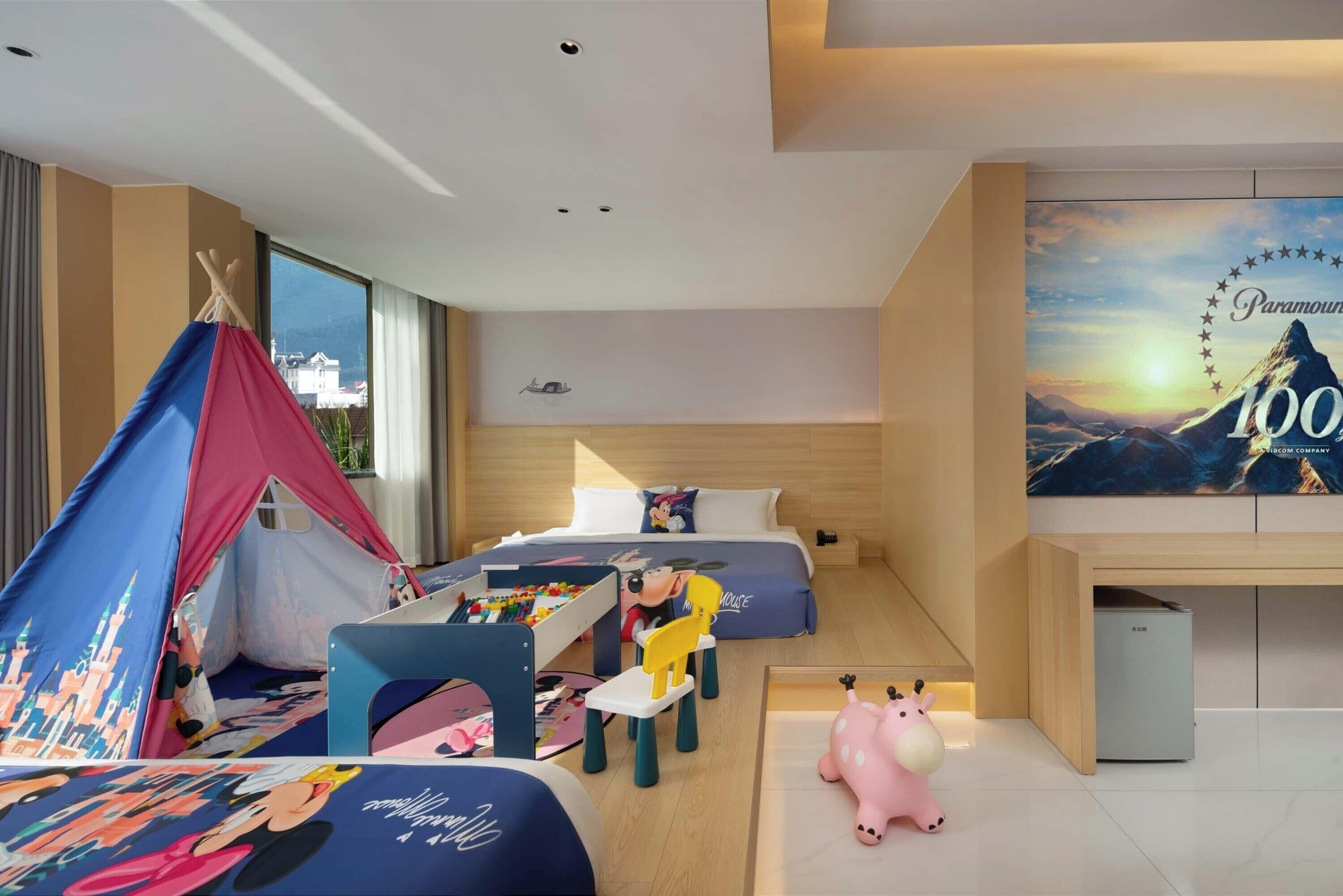 Une chambre d’hôtel moderne avec un grand lit, un petit bureau, un espace de jeu aménagé comme une tente et une grande affiche au mur.