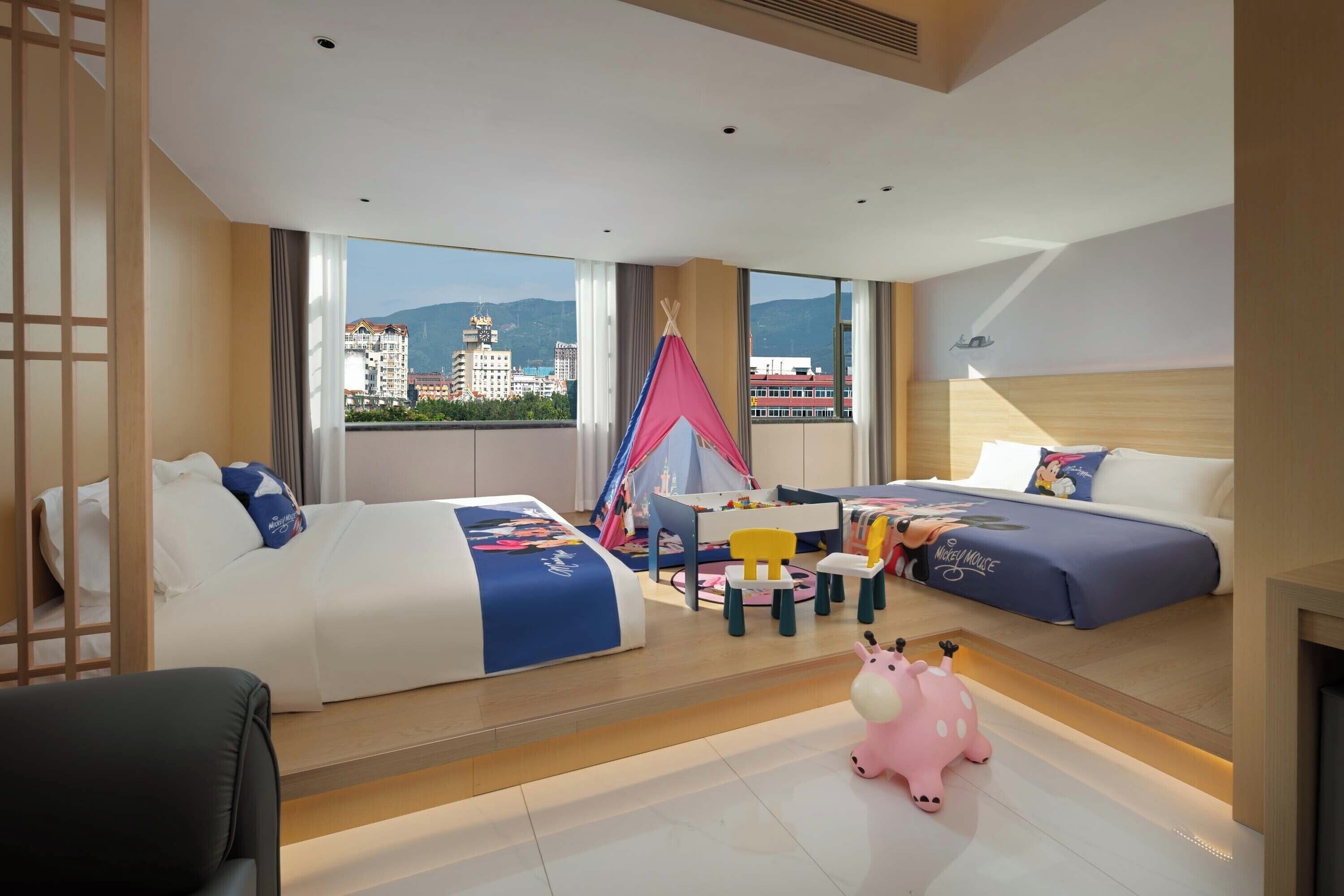 A modern hotel room with two beds, a small table, chairs, a pink toy, and a view of buildings and mountains.
