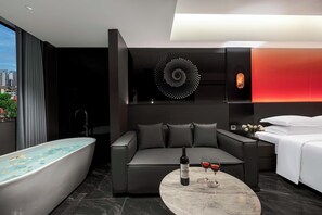 Una habitación de hotel moderna con una bañera grande, un sofá gris, una mesa redonda de mármol con una botella y copas, y vistas a la ciudad a través de ventanas amplias.
