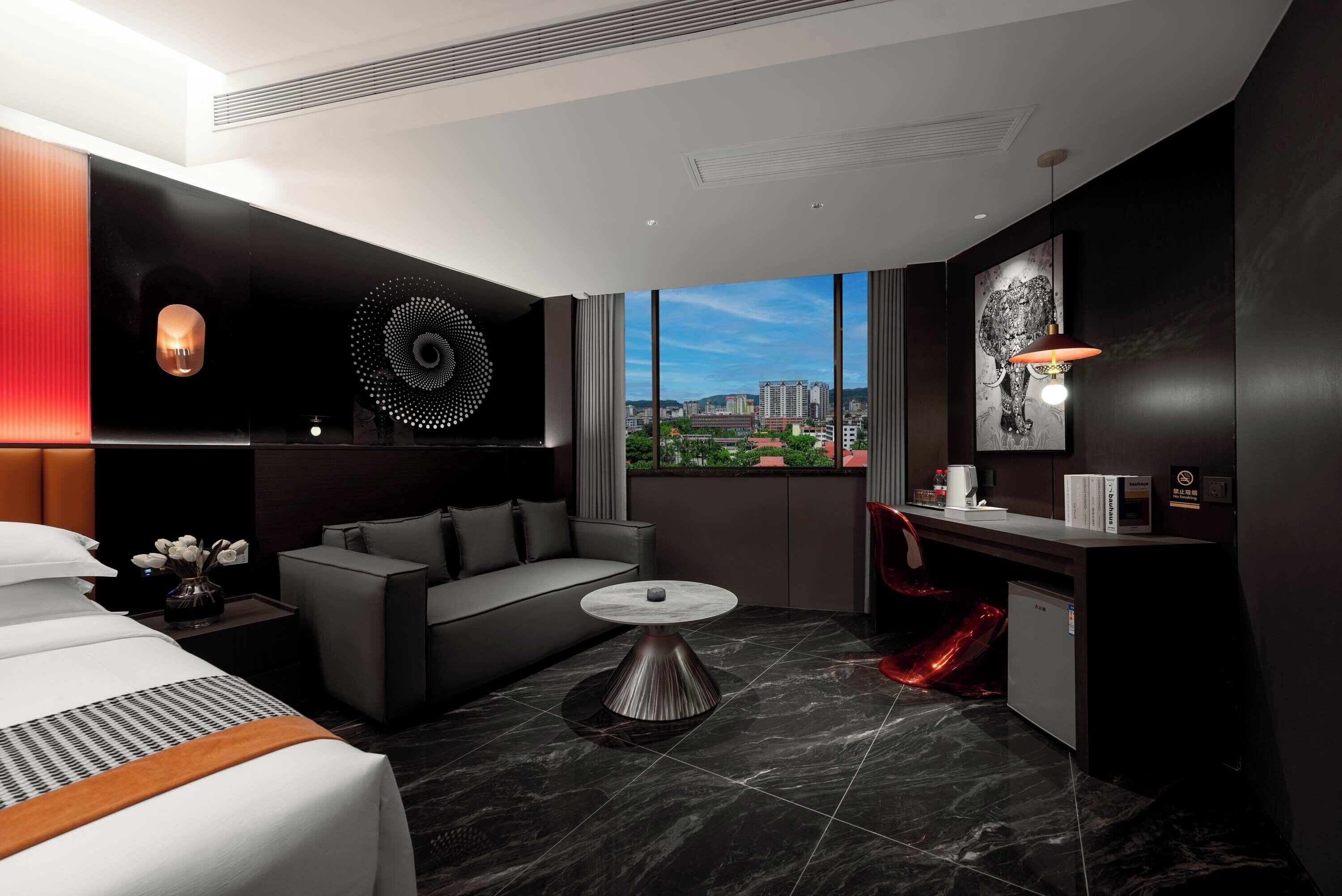 A modern hotel room with a bed, a sofa, a small table, and a view of the cityscape through large windows.