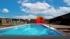 Una piscina en la azotea con un mural de atardecer, sillas de descanso y vistas de la ciudad.