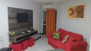 A living room with a red sofa, a TV mounted on a wooden wall, a small table with a phone and a vase, and a red chair. - Apartment for up to 6 adults, 3 minutes from Hot Park, simple and cozy (Rio Quente)