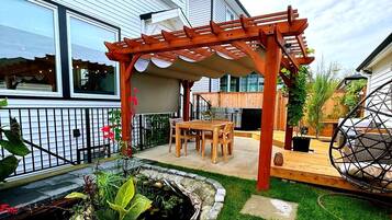 A backyard with a wooden pergola, a dining area with chairs, a small pond, and a garden with various plants.
