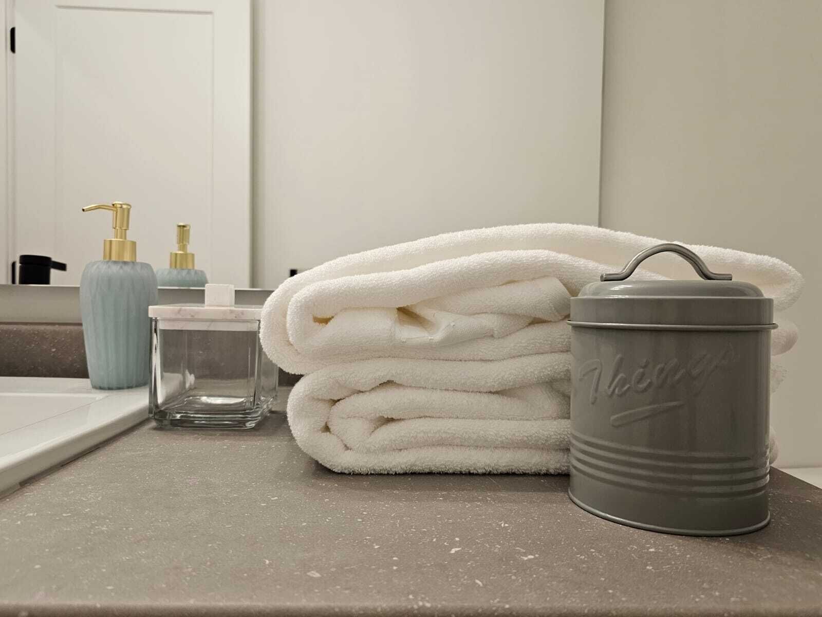 A bathroom with a sink, a towel dispenser, a glass container, and a grey tumbler on the counter.