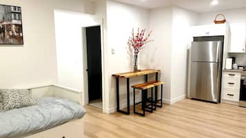 A compact living space with a sofa, a wooden table with benches, a refrigerator, and a kitchen area.