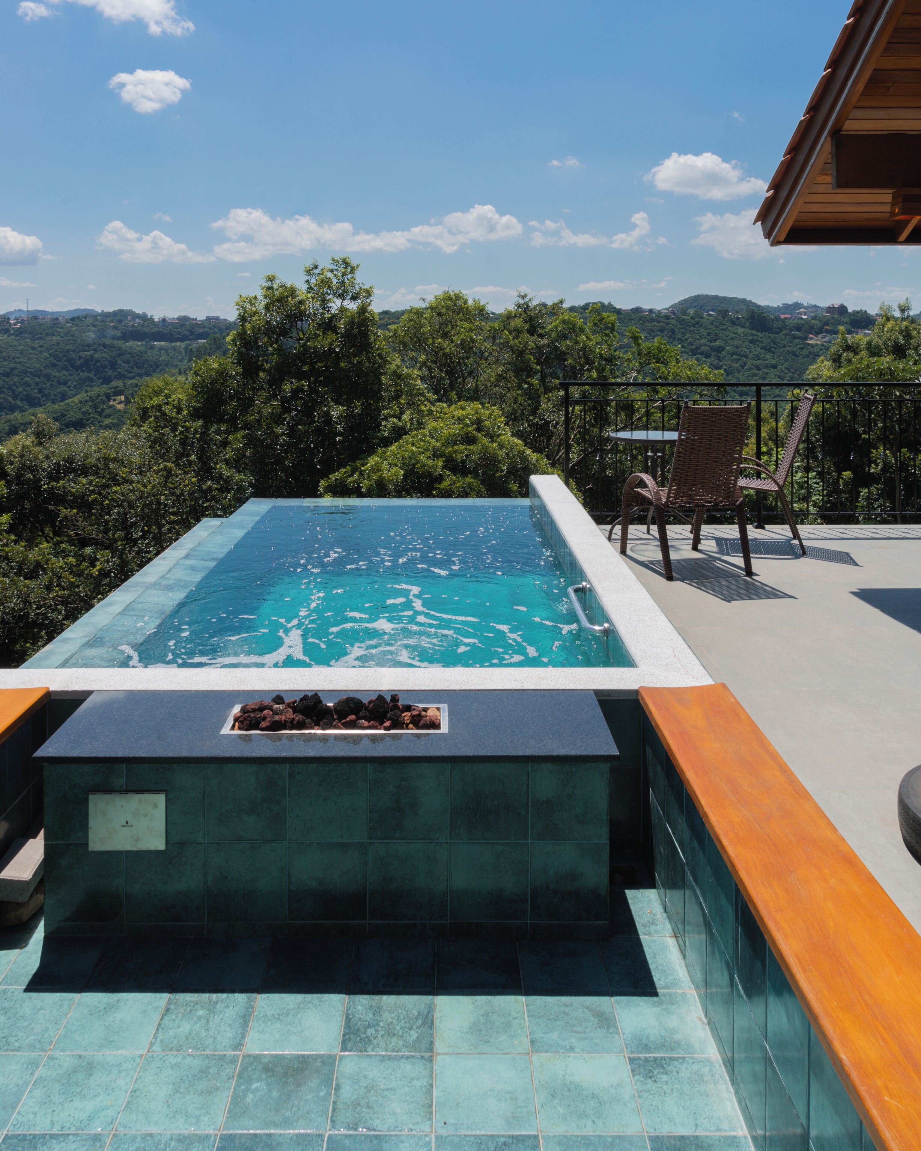 A rectangular infinity pool with a wooden deck and a chair, surrounded by lush greenery and a mountainous backdrop.