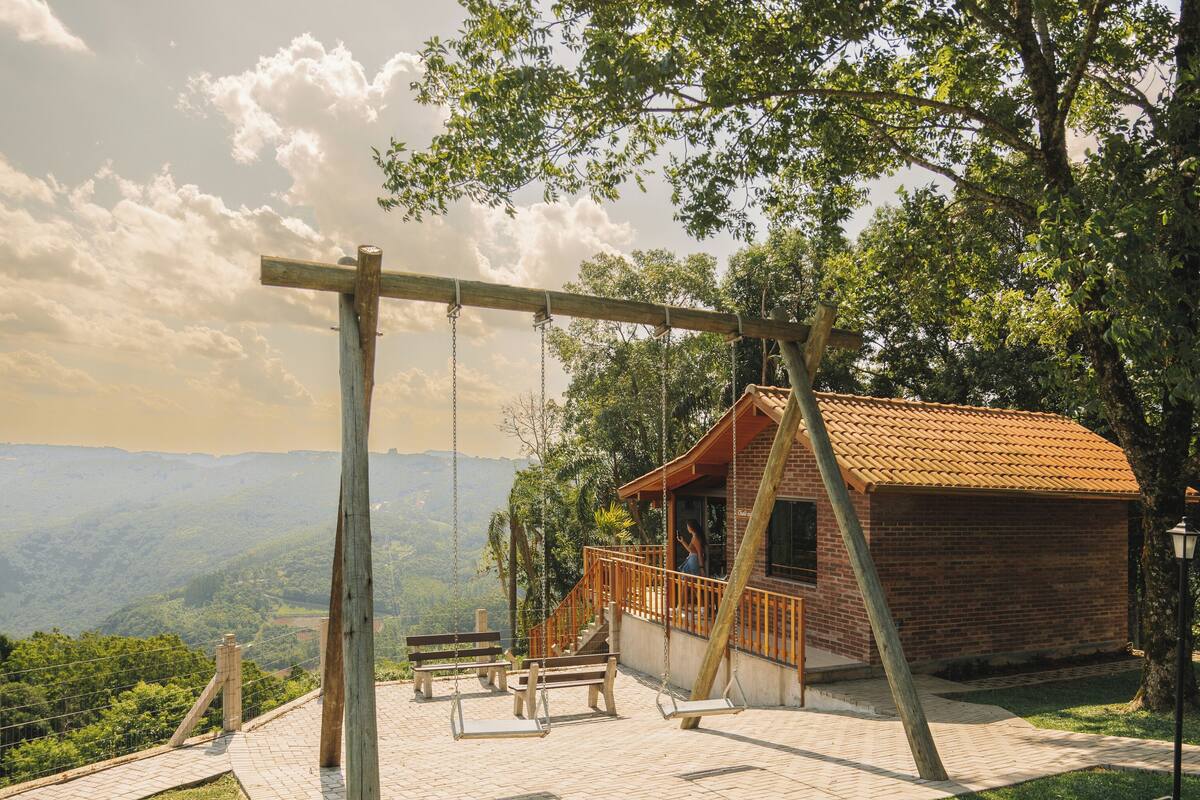 Una cabaña rústica con un columpio y mesas de picnic en un área verde, con vistas a montañas distantes.