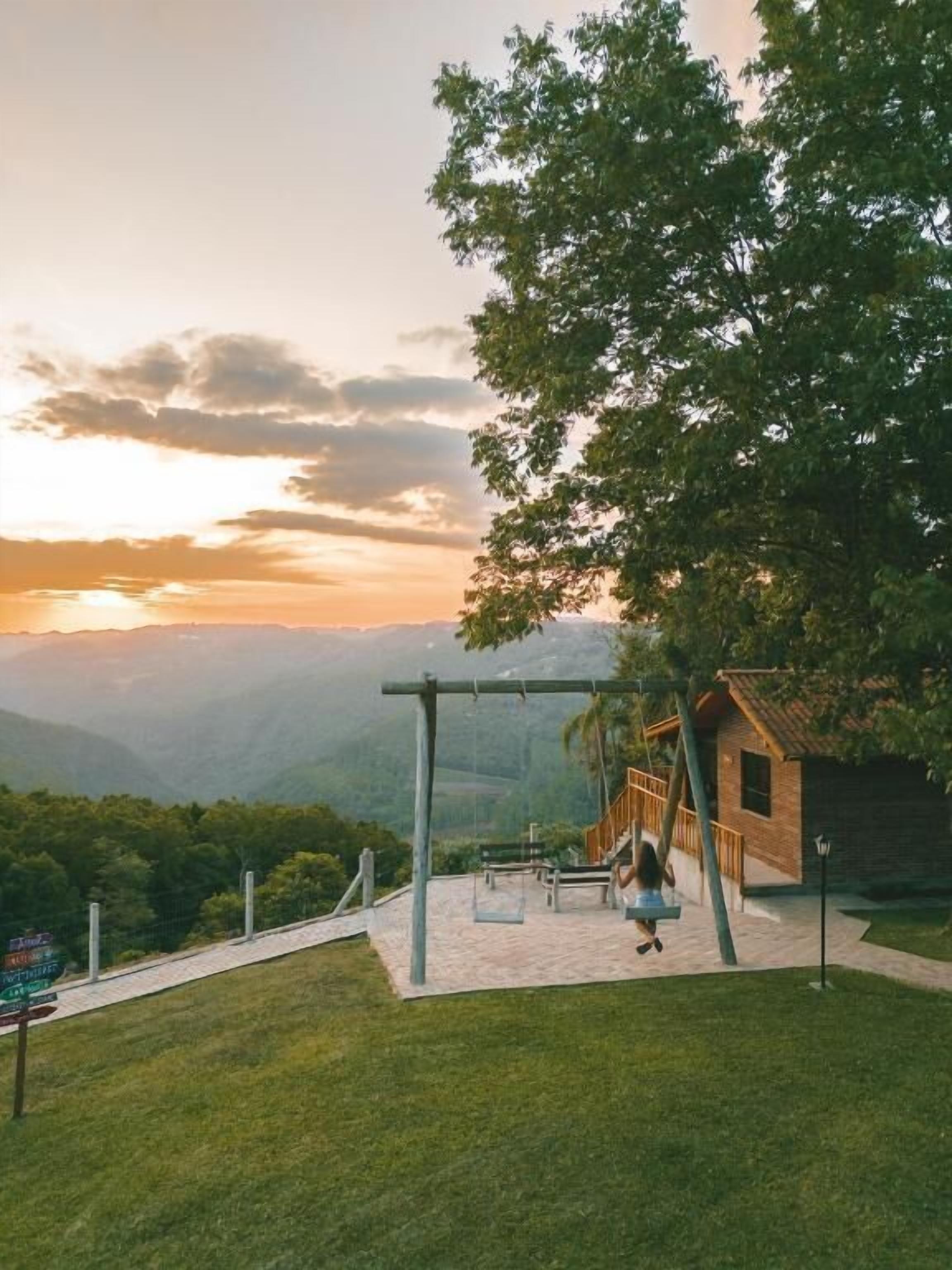 A park with a swing set and a small wooden building, surrounded by lush greenery and a mountainous backdrop.