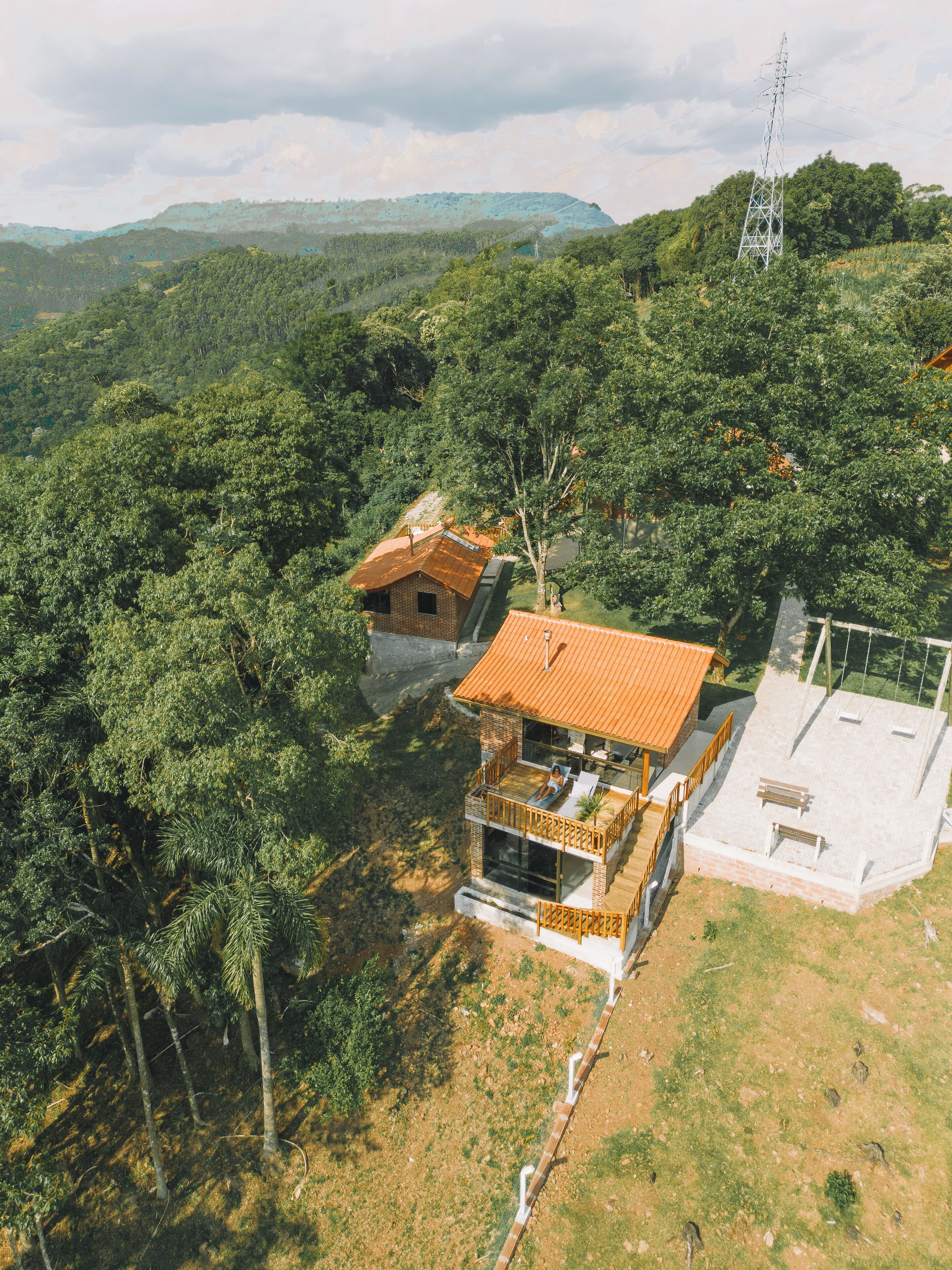 A modern house with a large deck and a separate building with a flat roof, surrounded by dense forest and hills.