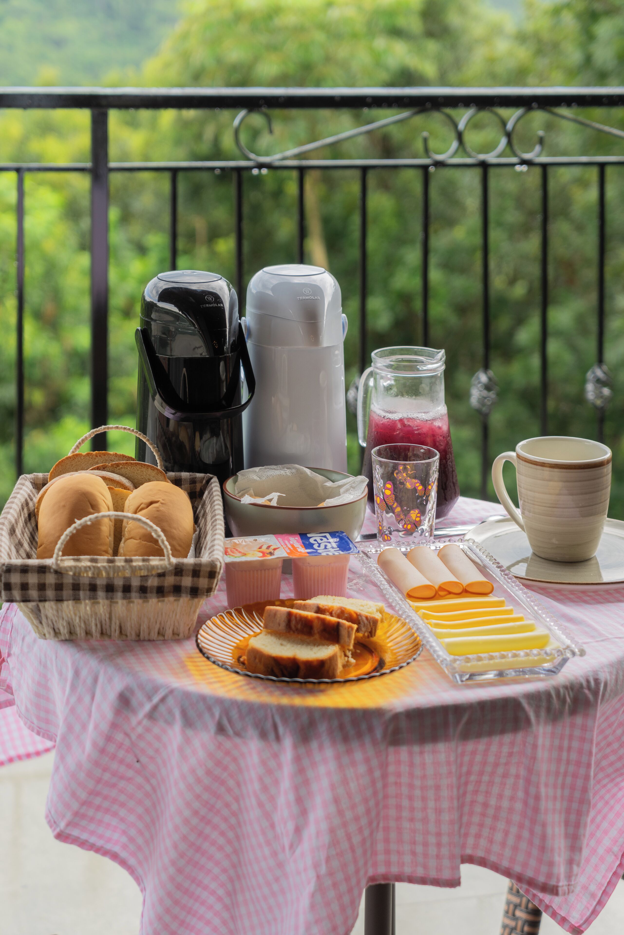 En frukostuppsättning på en balkong med en kaffebryggare, en kanna, ett glas juice, en mugg, en korg med bröd och en tallrik med bakverk.