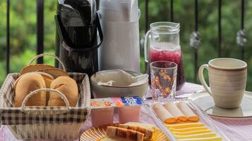 En frukostuppsättning på en balkong med en kaffebryggare, en kanna, ett glas juice, en mugg, en korg med bröd och en tallrik med bakverk.
