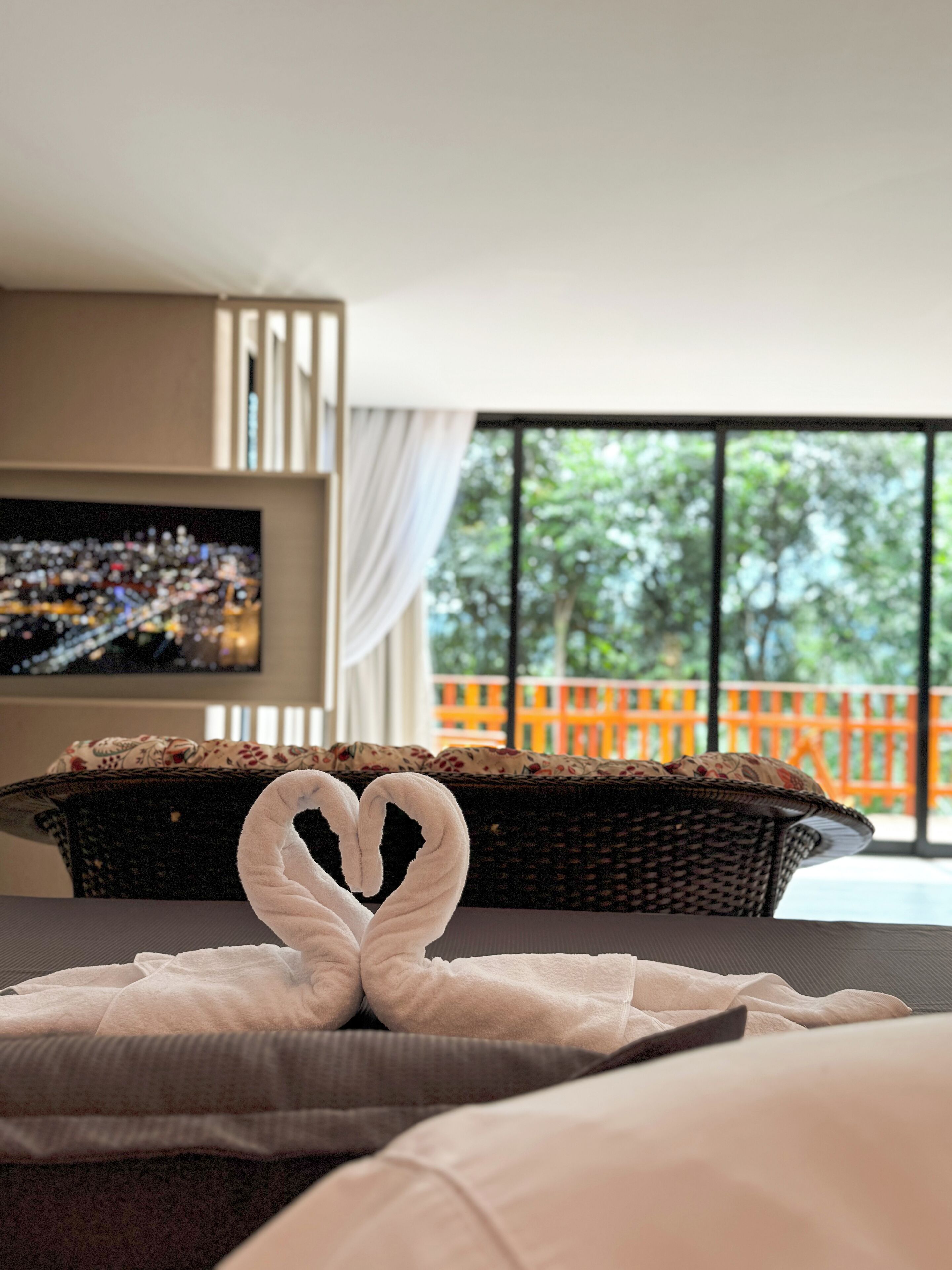 A hotel room with a large window, a television, and a heart-shaped towel on the bed.