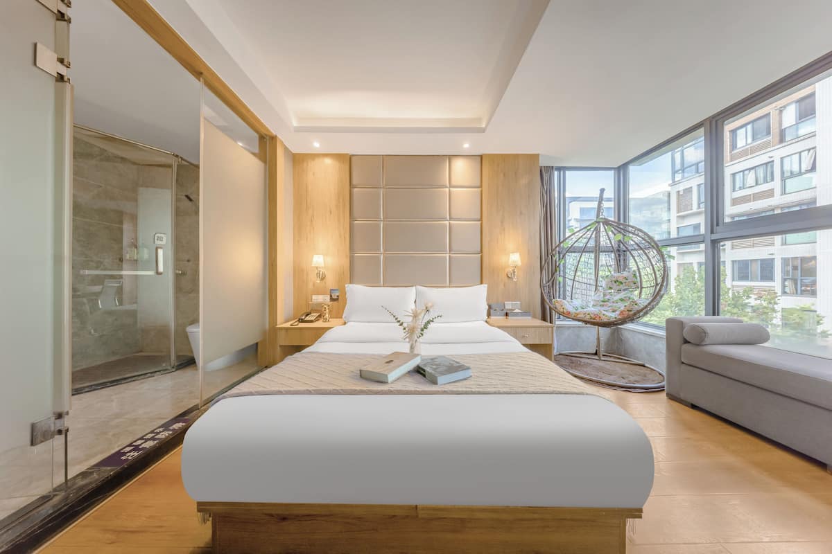 A modern bedroom with a large bed, a glass shower enclosure, a seating area with a cushioned bench, and a view of a building through large windows.