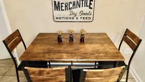 A wooden dining table with three candles, surrounded by wooden chairs. A sign on the wall reads "Hardware Mercantile Dry Goods Notions & Feed."