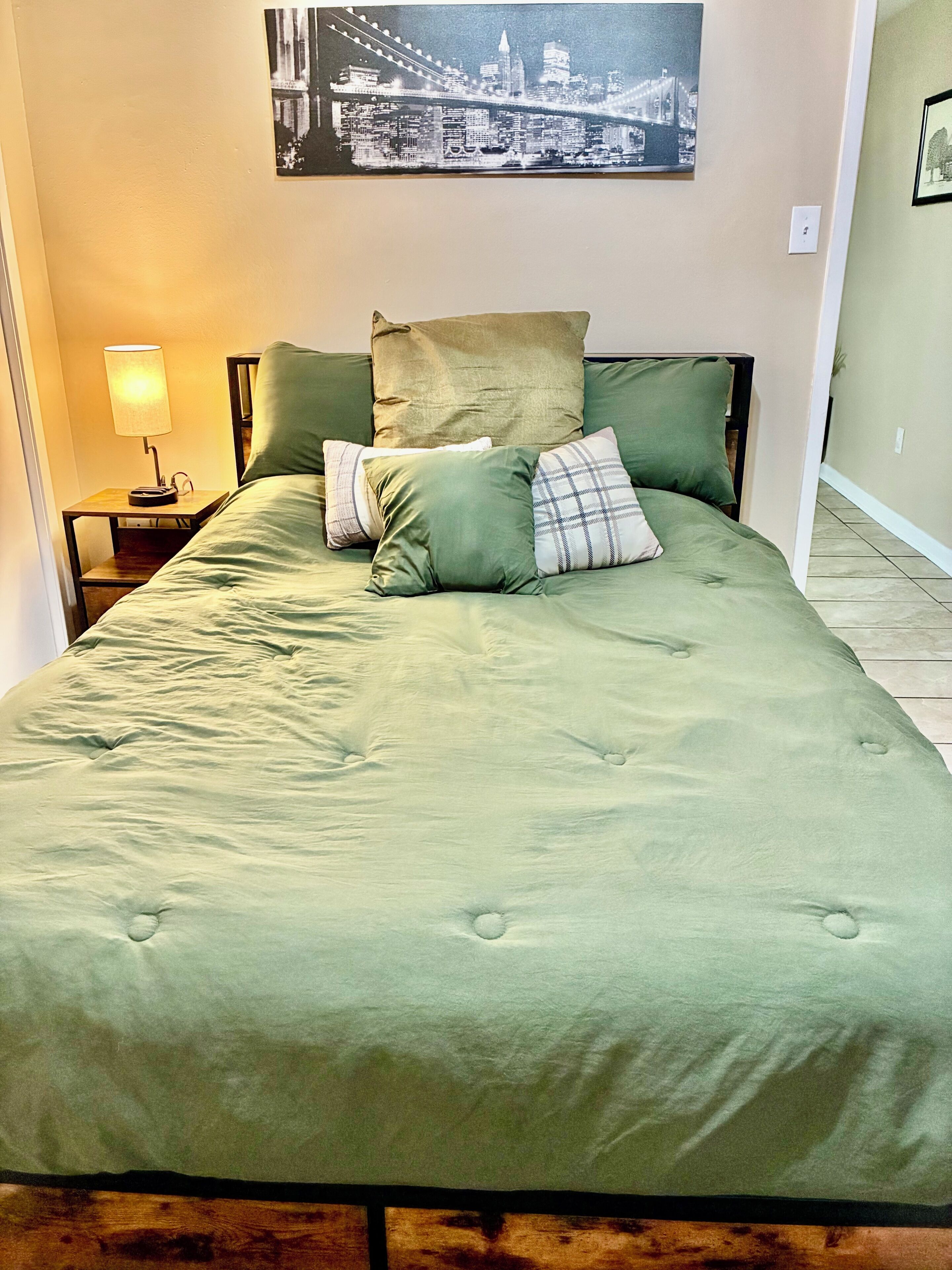 A bed with a green bedspread and multiple pillows, a nightstand with a lamp, and a cityscape picture on the wall.