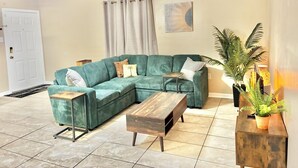 A living room with a sectional sofa, a wooden coffee table, a ceiling fan, a door, and a potted plant.