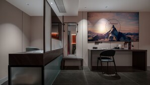 Ein modernes Hotelzimmer mit Schreibtisch, einer Sitzbank und einem großen, an der Wand montierten Fernseher, der einen Sonnenuntergang über Bergen zeigt.