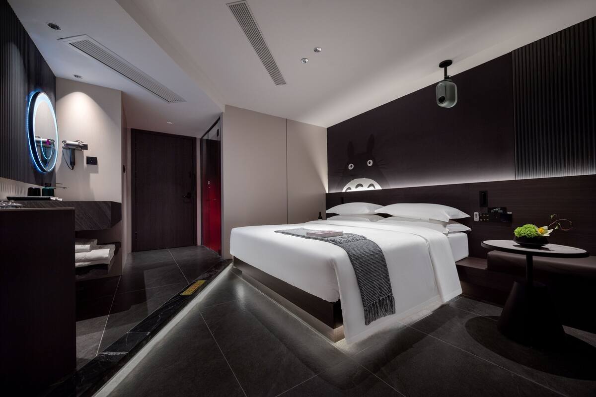 A modern hotel room with a large bed, a bedside table, a mirror, and a wall-mounted camera.