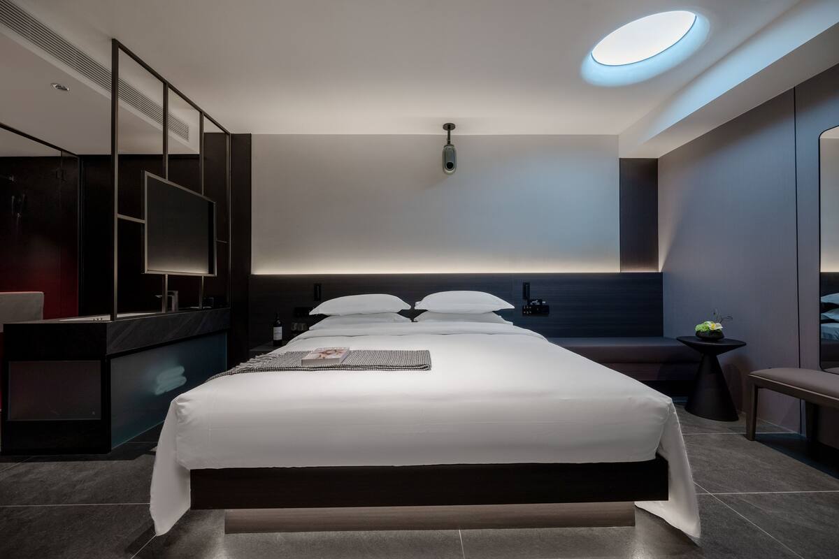 A modern hotel room with a large bed, a bedside table, a small side table, a built-in TV, and a ceiling light.
