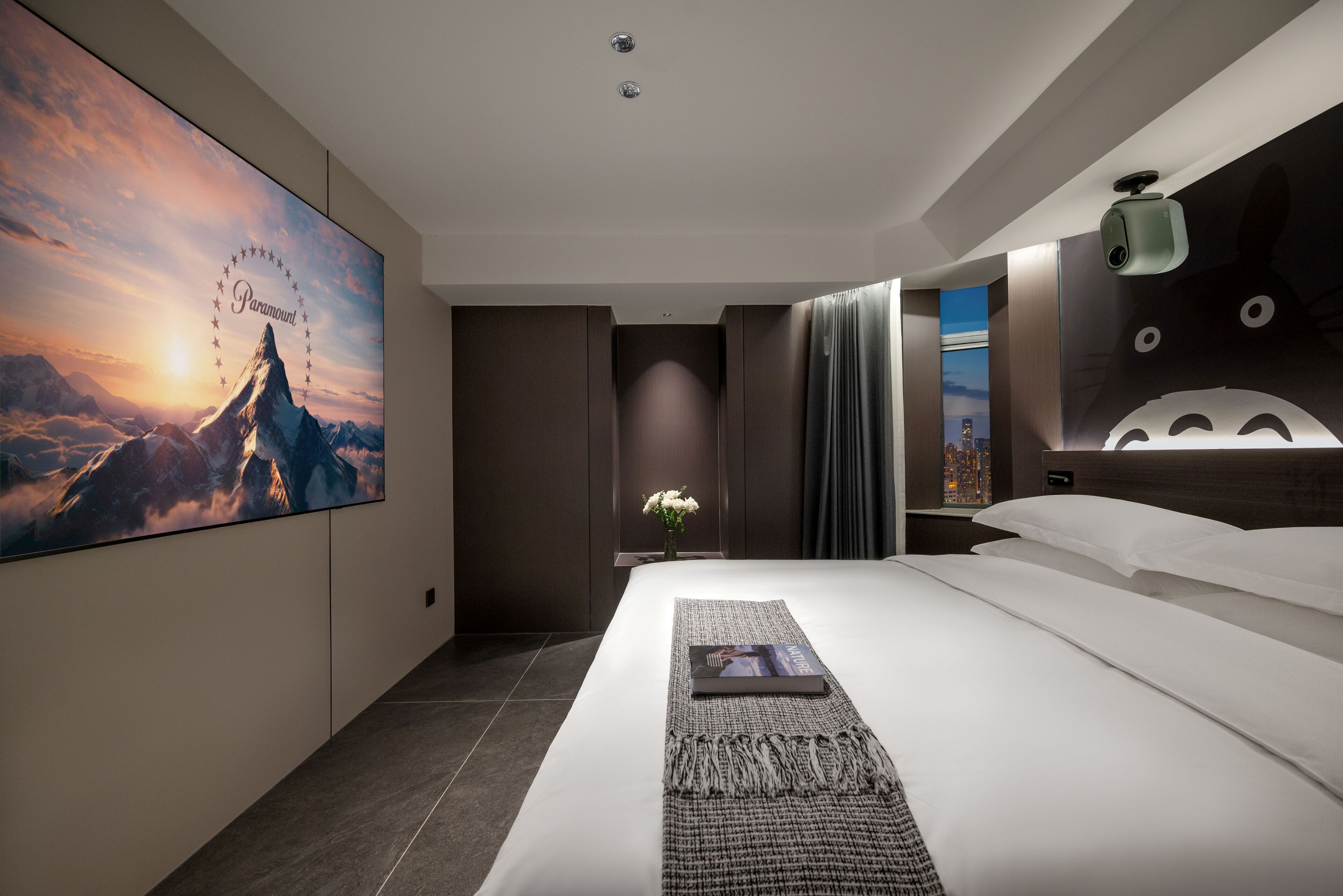 Een moderne hotelkamer met een groot panoramisch wanddecor, een netjes opgemaakt bed, een nachtkastje met een lamp en uitzicht op de stad door het raam.